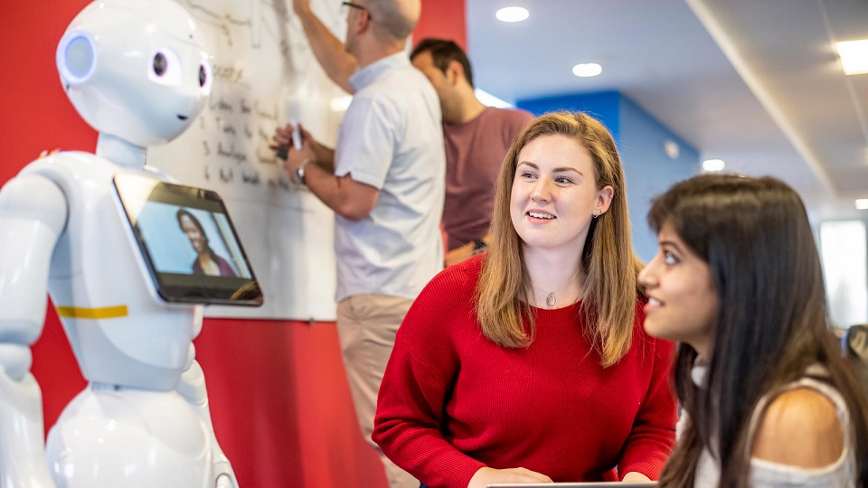 Two female students who are sitting down facing humanoid robot pepper.