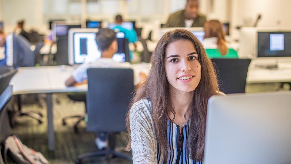 Female student sat at a computer, with several other students blurred in the background in the computer room.