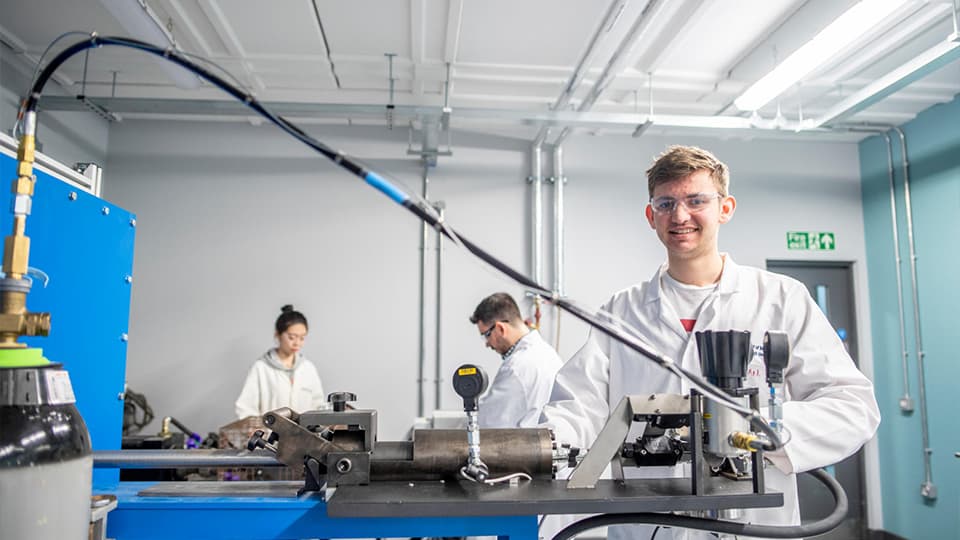 student in lab with equipment smiling at camera and 2 other students in the background