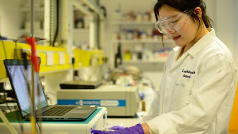 female in lab coat and goggles with laptop on benchtop in a lab