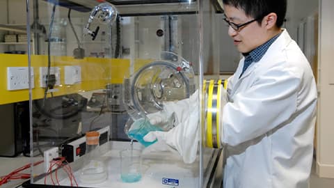 male in lab coat pouring blue liquid from a beaker into another beaker in an isolated cabinet