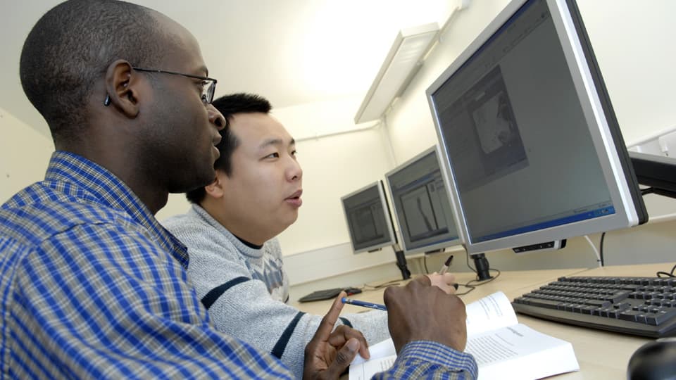 two students using computers with books spread out on the desk