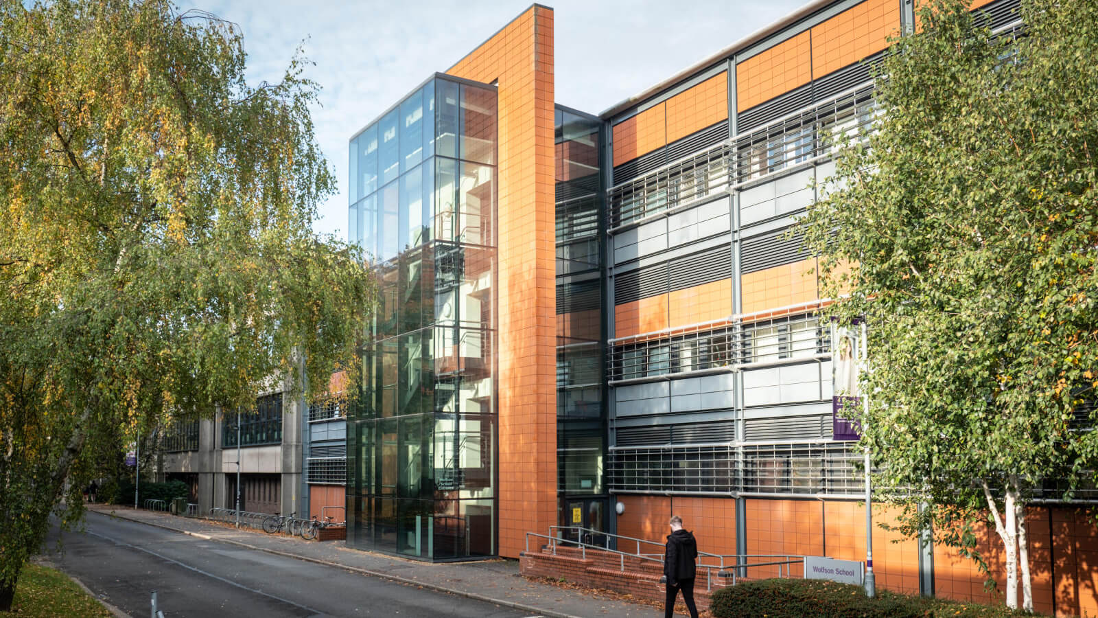 Wide shot of the outside of the Wolfson School of Engineering with a student walking past in the opposite direction to the camera.