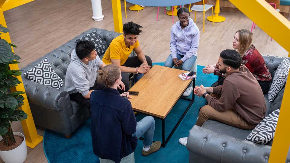6 students sitting around a table chatting and laughing in the student union. 