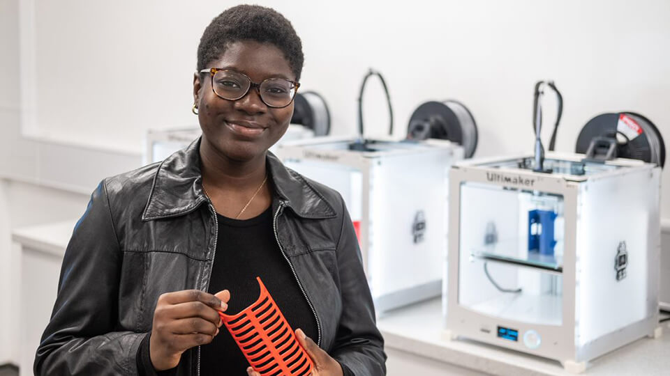 Image of Product Design Engineering student Tami smiling at the camera whilst holding a piece of equipment produced by the 3D printers which are featured in the background.