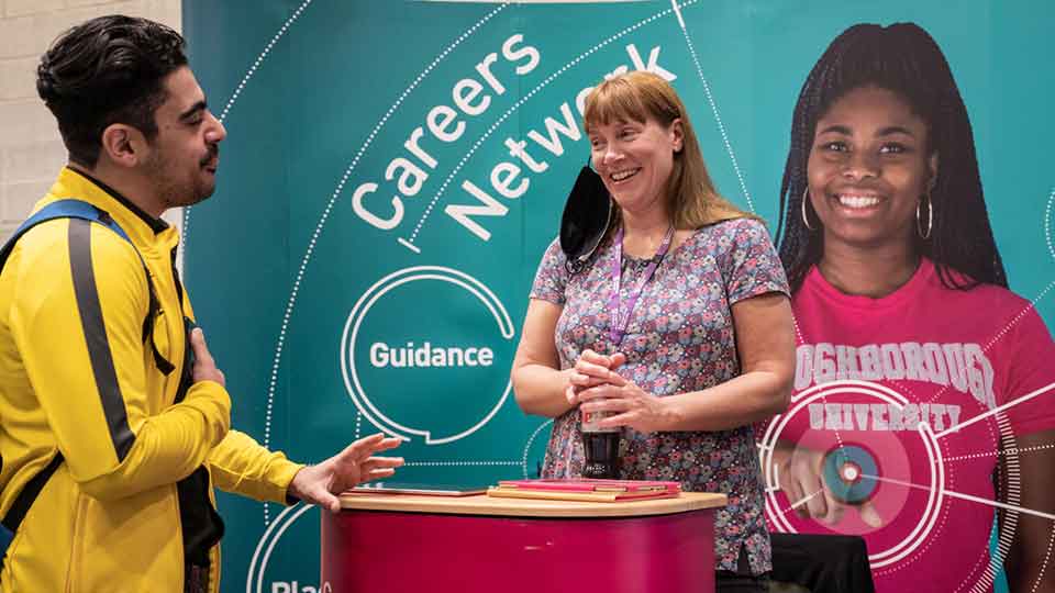 A female staff member stands at a promotional stand with a large banner showing 'careers fair' text. The female is engaging with a male while smiling and having a conversation. 