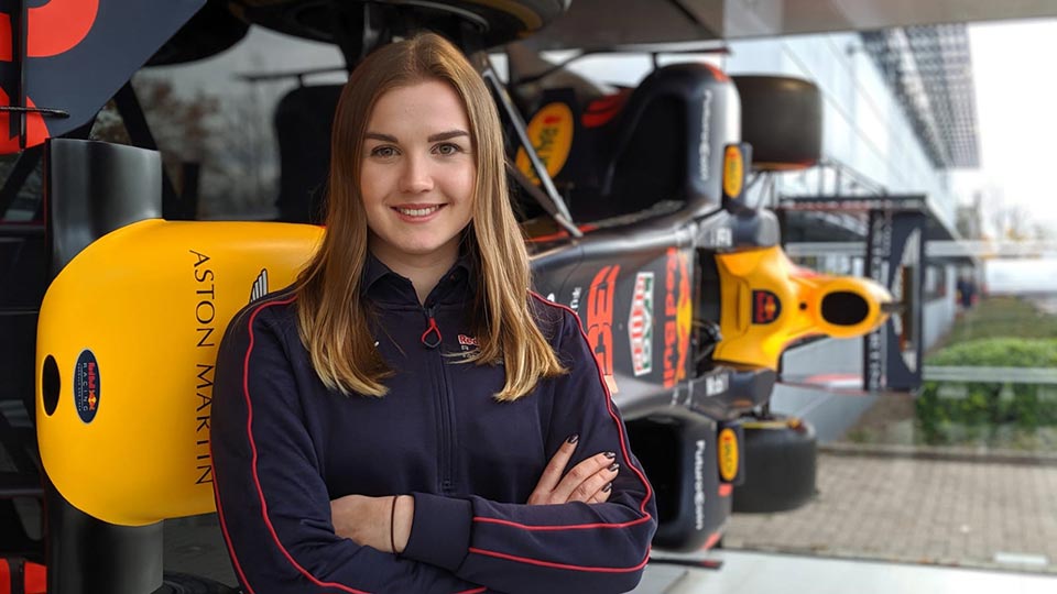 Headshot of graduate, Aimee Gibbard, standing with her arms folding whilst smiling, wearing a Red Bull racing jacket featuring a Red Bull Aston Martin racing car in the background.