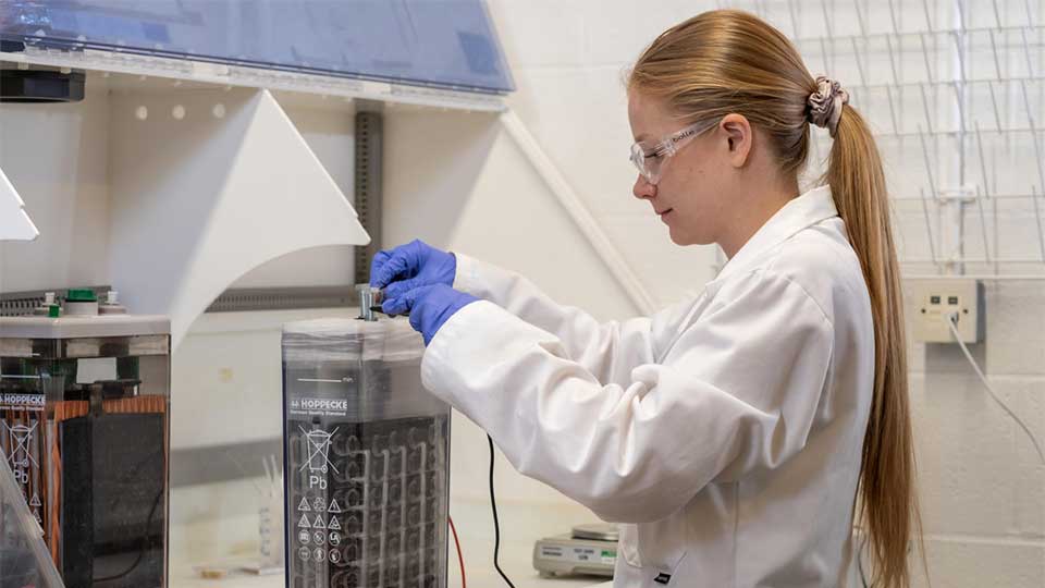 A student wearing goggles and lab coat, testing a battery in the lab.