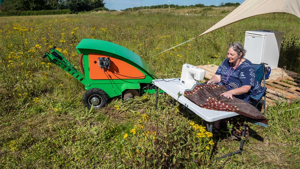 An academic sat at a table in a field using a sewing machine which is operated by a solar powered machine.