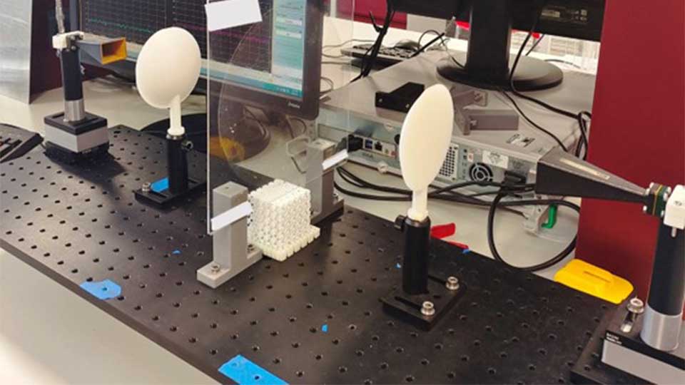 A lab bench setup where a transparent rectangle in the middle of the setup that contains a circle of a different transparent material. Approximately 10 centimetres on either side of the transparent sheet are two 3D printed lenses that resemble flattened half-ellipsoids.