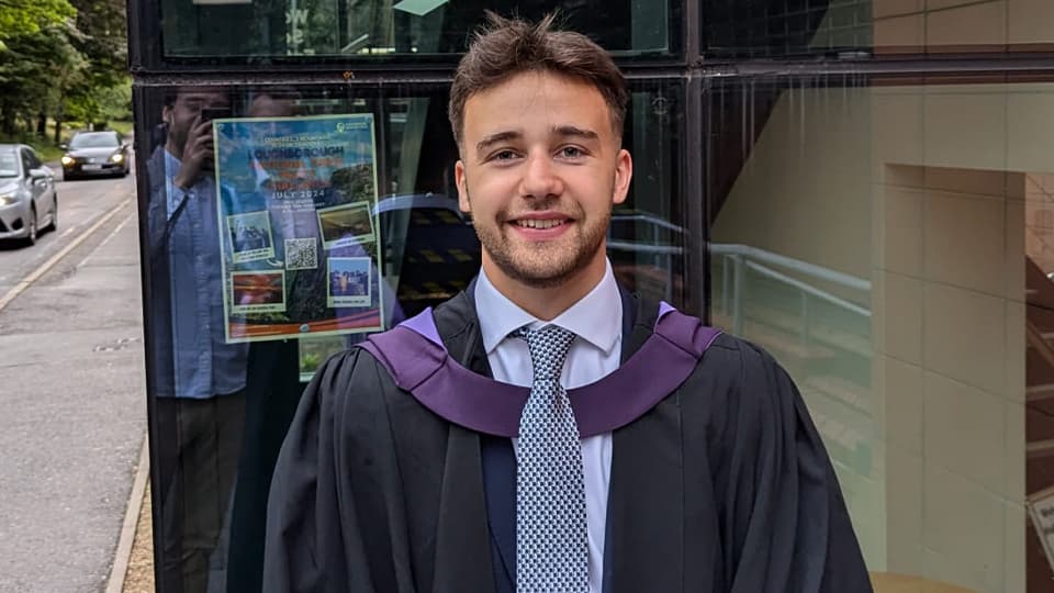 Adam stood in graduation gown by a University building