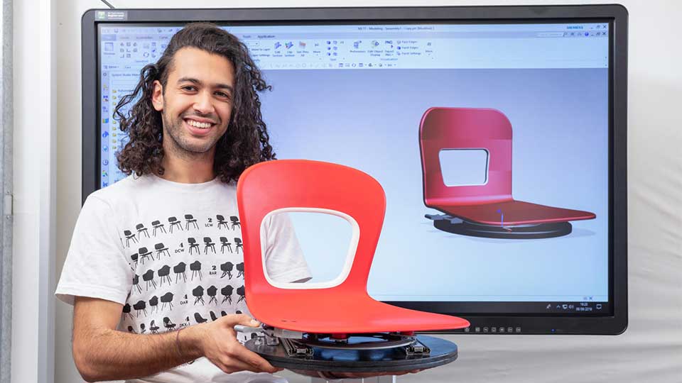 A student sat in front of an interactive screen which shows a CAD model of a red chair. The student is holding a red chair whilst smiling and looking directly at the camera. 