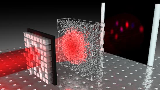 A scientific visualization of a laser beam passing through different optical elements. The image shows a structured light source emitting a red laser beam that passes through a complex, translucent medium with scattered particles. The beam then reaches a screen, where a diffraction or interference pattern is displayed. The setup appears to be part of an experiment in wavefront shaping, light scatt