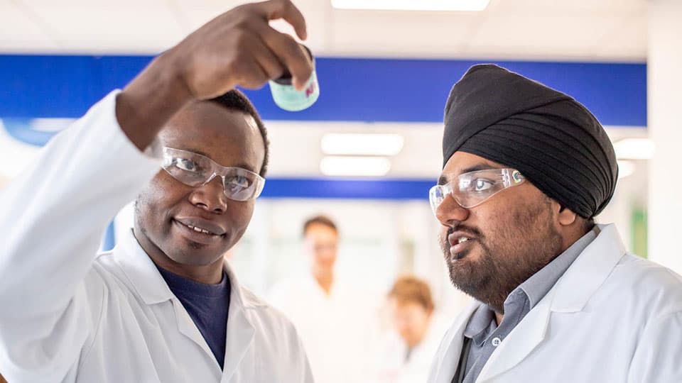 Two students in a laboratory wearing white lab coats and goggles, inspecting the contents of a container.