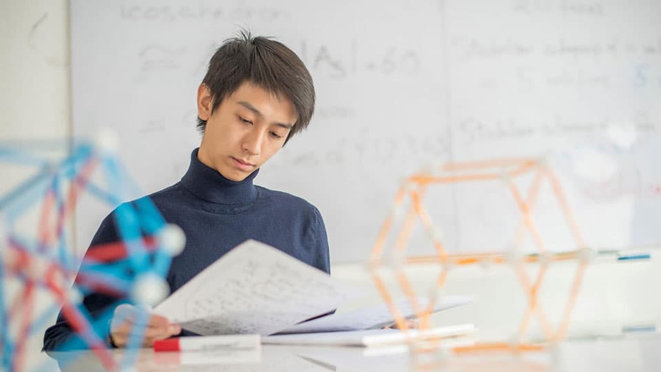 A student in the Mathematical Sciences department looking at paper-based work with physical mathematical objects on the table in front.