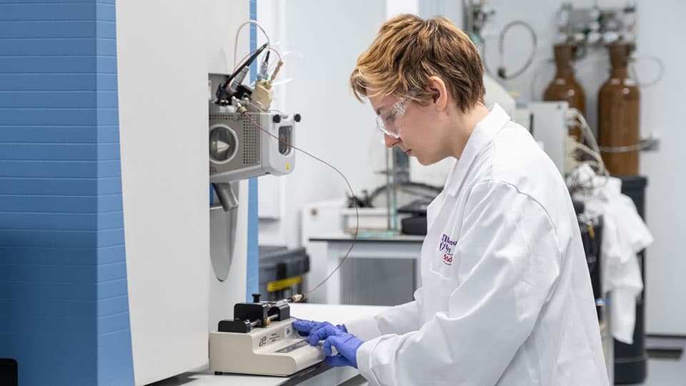 Someone in a white lab coat and goggles working in an analytical laboratory