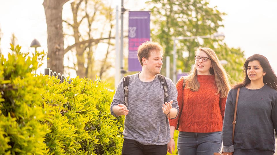 Three students walking outside with green trees and bushes behind them