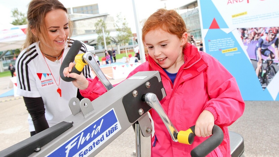 A female child is shown how to use a hand exercise bike by one of the volunteers at a National Paralympic Day event