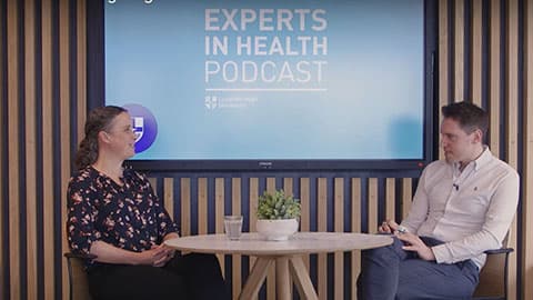 A man and woman sat at a table with the words Experts in Health podcast on the wall behind them