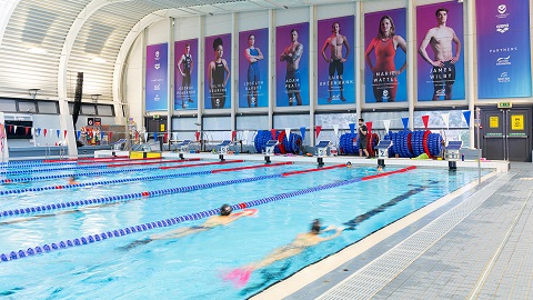 The Olympic-sized swimming pool at Loughborough University. Swimmers are training in the pool. There are posters displayed of Team GB swimmers.