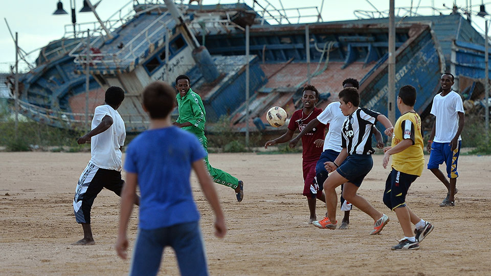 Children playing football in front of a rusting ship