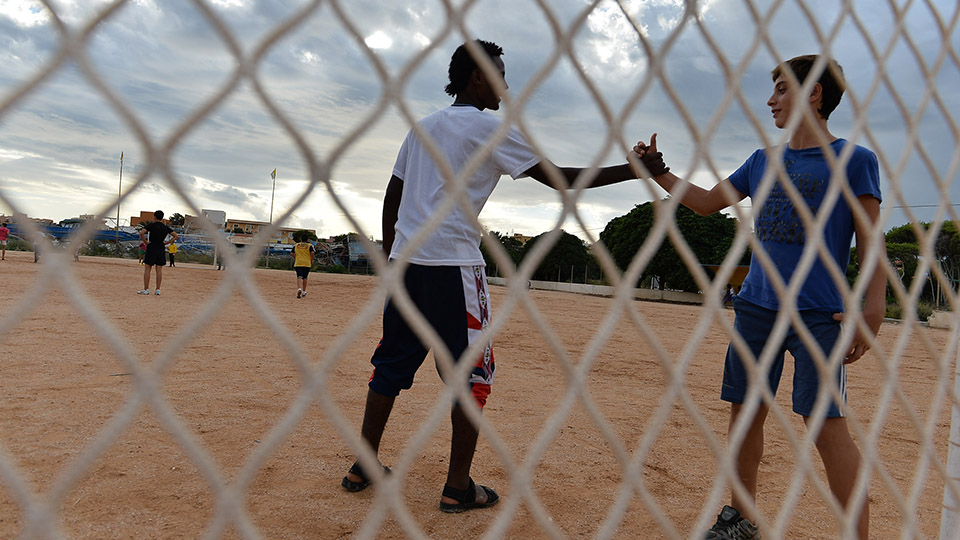 Two young men shaking hands behind a mesh fence. Photo credit: Tullio Puglia