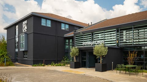 Modern exterior of Burleigh Court Hotel featuring dark grey walls, large glass panels at the entrance, potted trees, and outdoor seating under a partly cloudy sky.