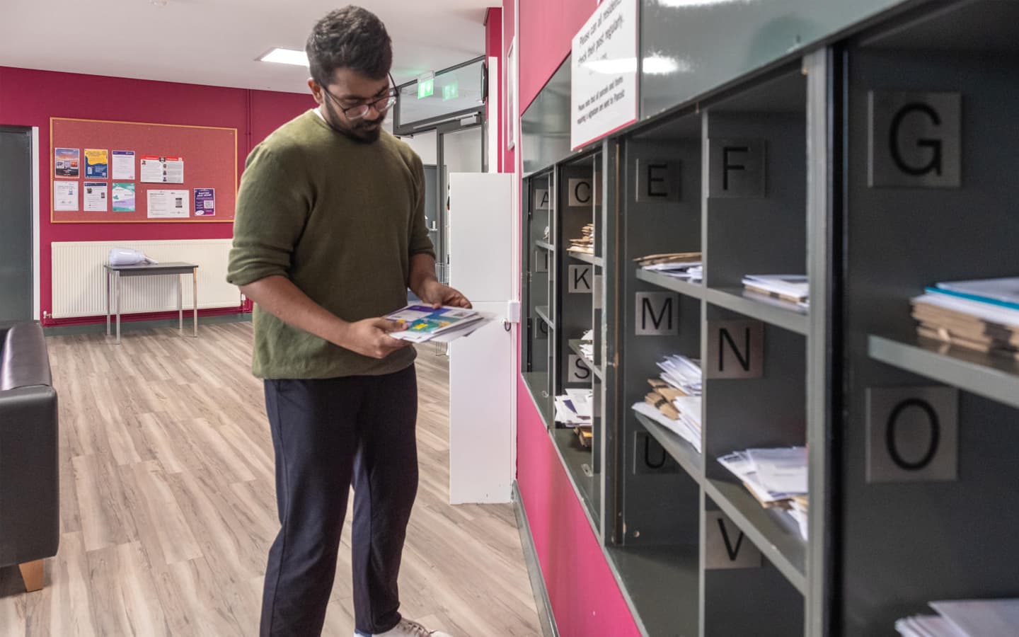 A person sorting papers in front of alphabetically labeled mailboxes in a hallway with red walls and wooden flooring. A bulletin board with notices and a table with a chair are visible in the background
