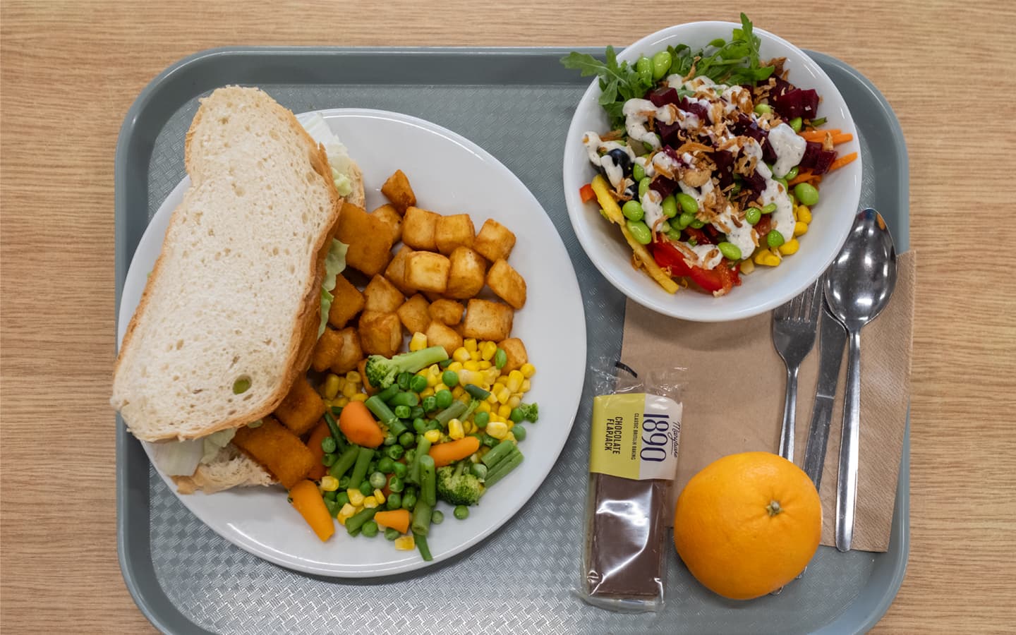 A cafeteria tray with a balanced meal: a plate containing a large sandwich with white bread, golden-brown roasted potato cubes, and mixed vegetables including carrots, broccoli, green beans, and sweetcorn. Next to the plate is a bowl of colorful salad topped with dressing and seeds, a wrapped chocolate brownie labeled ‘1890’, and a whole orange. A fork and spoon rest on a napkin on the tray
