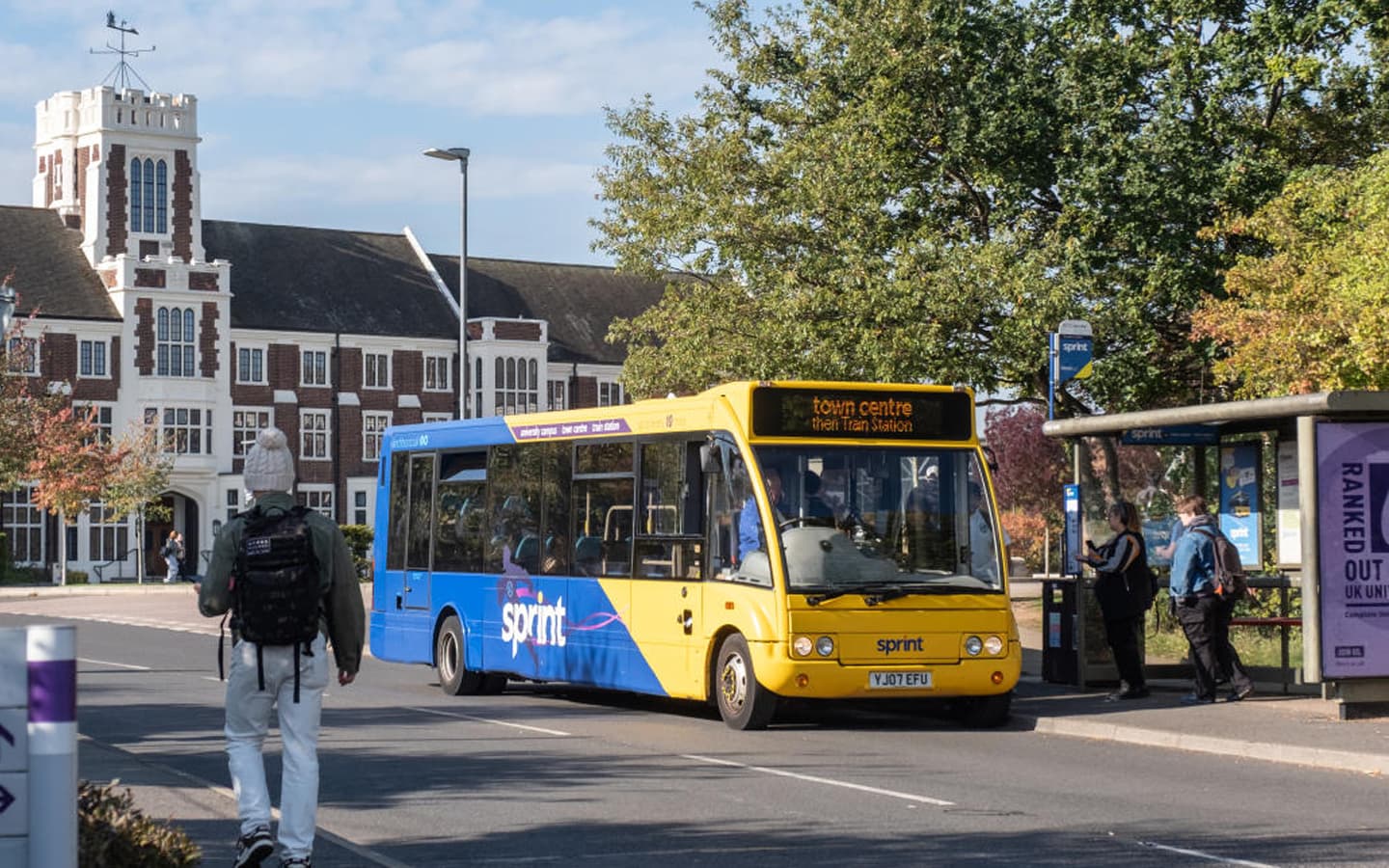 A blue and yellow Sprint bus at a bus stop with a sign showing destinations including town centre and train station. Several people are near the stop, and a person with a backpack is walking along the road. In the background, there is a large historic-style building with white and red brickwork, trees with autumn foliage, and a clear blue sky