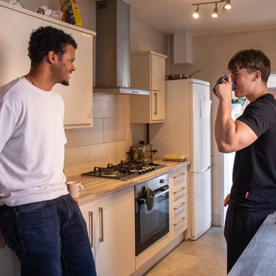 A modern shared kitchen with light-colored cabinets, a built‑in oven, a gas hob, and a tall fridge‑freezer. Two people stand talking near the countertop, with one leaning against it and the other holding a mug. Warm overhead lighting brightens the room.