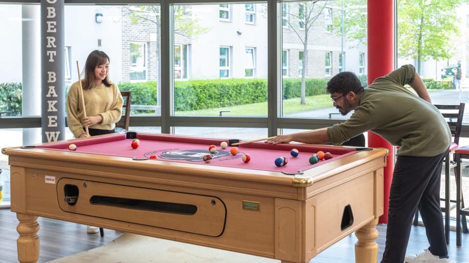 Two people playing a game of pool on a red pool table in a bright common room with large windows overlooking a courtyard.