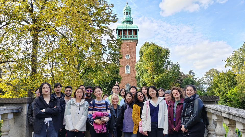 A group of international students stand facing the camera smiling in Queen's Park Loughborough in front of the Carillon bell tower