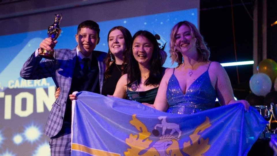 A group of students standing together on a stage holding a David Collett hall flag.