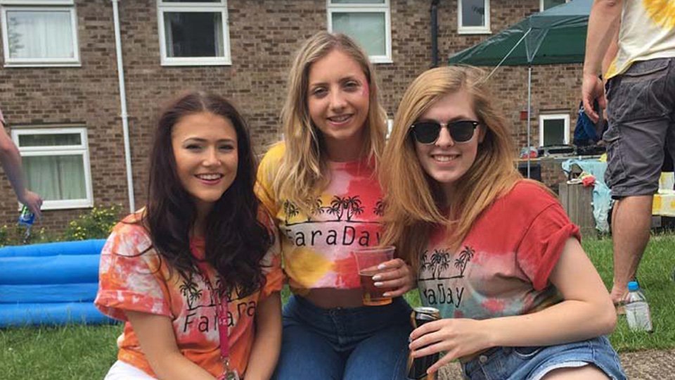 Three female students sitting outside on the grass outside Faraday hall. They are wearing brightly coloured Faraday hall t-shirts and smiling.