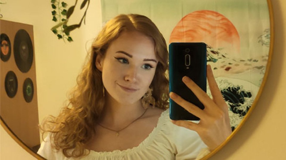 A female student taking a selfie in a hanging mirror in a Royce hall bedroom. She has art posters hung up on the walls behind her.