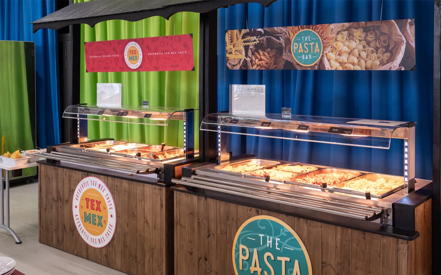 “Two self-service food counters in a dining area, labeled ‘Tex Mex’ and ‘The Pasta Bar.’ Each counter has a wooden base with signage and a heated display case containing trays of hot food. Colorful banners above the counters show pasta and Tex-Mex themes, and the background features bright green and blue curtains.