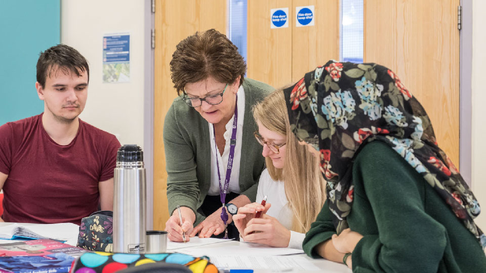 A student support staff member guides three students through paperwork on a shared desk.