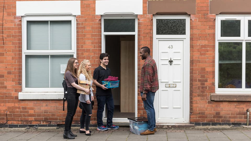 A group of four students stand in front of a town house in Loughborough centre, holding boxes ready to move in