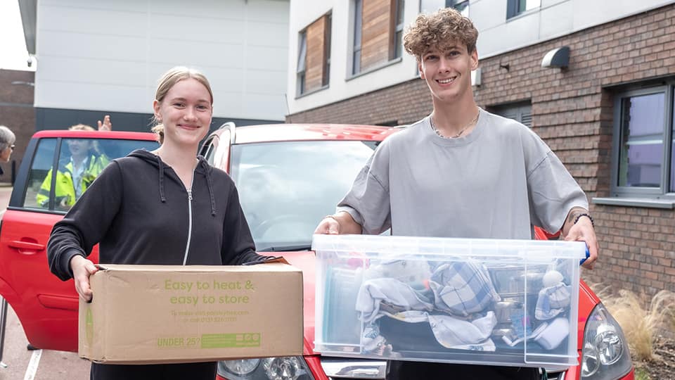 Two students smiling and holding boxes full of student items ready to move into their accommodation at Loughborough University