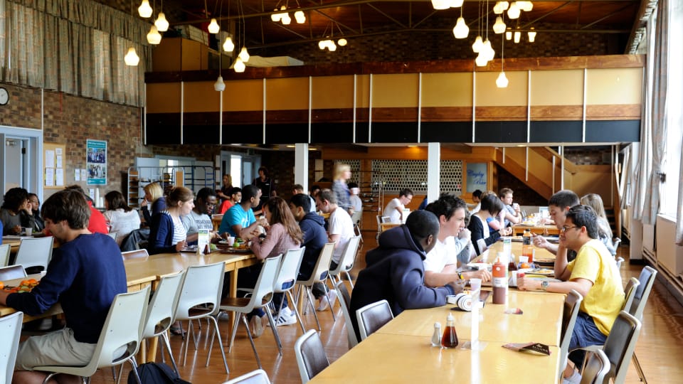 A busy dining hall with long wooden tables and rows of chairs, where many people are seated eating and talking. The room features warm overhead lighting, brick walls, large windows along one side, and an upper mezzanine level in the background.
