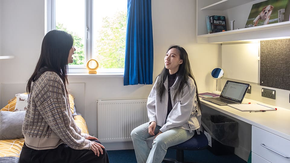 Students sat opposite each other inside a Falkner Eggington bedroom, one is sat at their desk chair and the other is sat on the bed, they are talking and laughing with each other