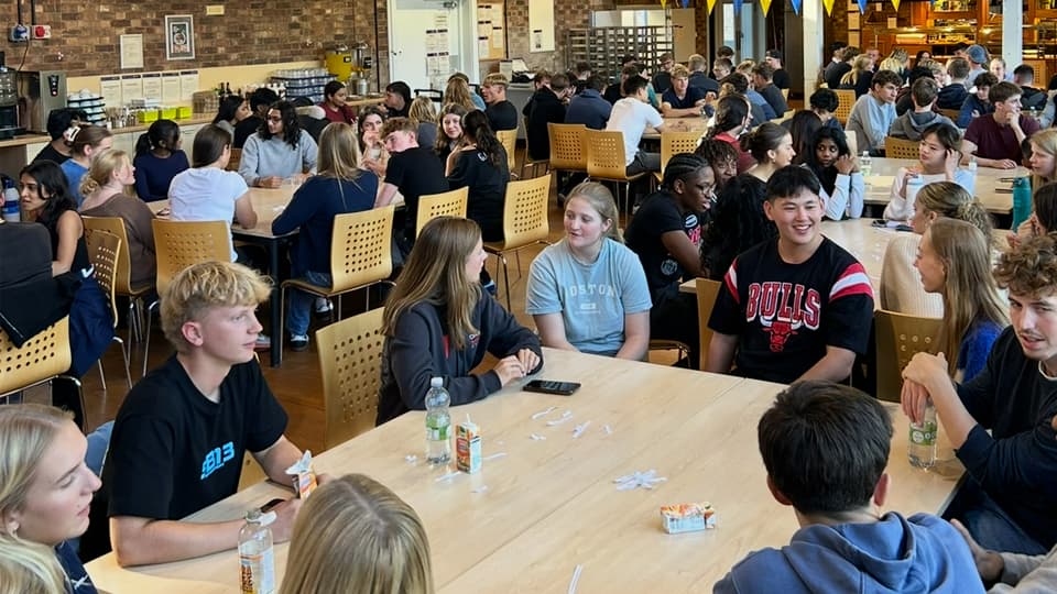 A large group of students sitting at tables in a dining hall or common area, talking and taking part in an icebreaker activity, with drinks and snacks on the tables.