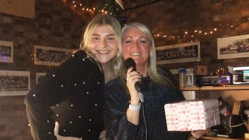 Two people standing together at an indoor event, one holding a wrapped gift and a microphone. The setting is warmly lit with string lights and framed photos on the wall behind them.