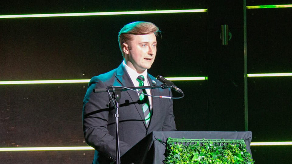 Student faces an audience of students at an awards night, he is stood speaking at a microphone and wears a smart suit.