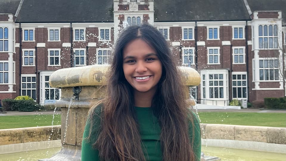 Loughborough University student sits on the iconic fountain outside the university's Hazlerigg building and smiles to the camera.