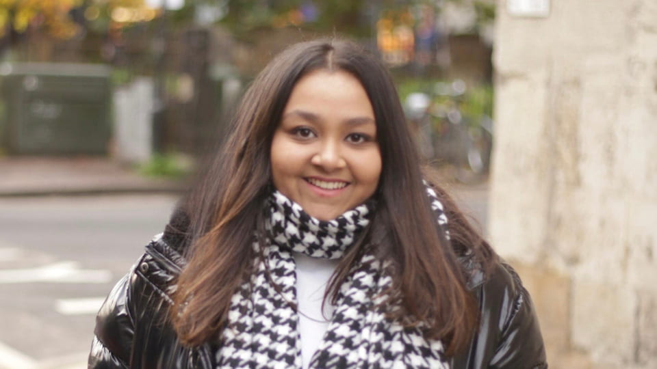 Student is wrapped up in warm autumn clothes, a thick black jacket and a checked scarf, and smiles to the camera