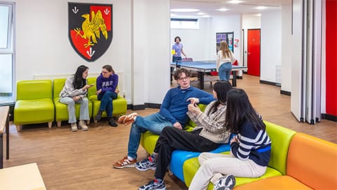 A group of 8 students in the common room sat chatting together on colourful chairs and playing a game of table tennis together.