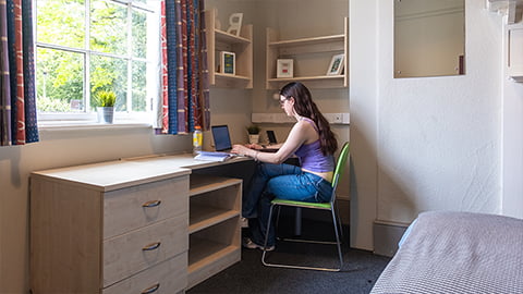 Student sat working at their desk in renovated Harry French bedroom with large study desk with a great view and a large bed