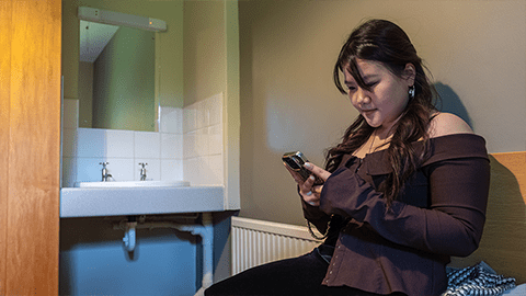 Student sat in Harry French bedroom next to her washbasin and is texting on her phone while sitting on her bed